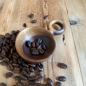 May include: A wooden coffee cup with a circular handle, filled with dark roasted coffee beans, rests on a rustic wooden surface. Scattered coffee beans surround the cup. The wood grain adds texture to the warm scene.