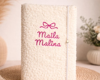May include: Cream-colored, fluffy journal with the embroidered name "Maila Malina" in pink script, topped with a bow design. A gray elastic band with a heart charm secures the journal. The journal is on a woven mat.