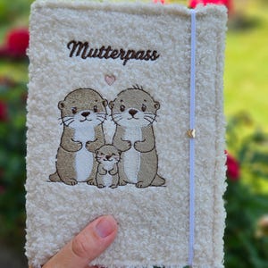 Könnte beinhalten: Eine cremefarbene Hülle aus strukturiertem Sherpa-Stoff für einen Mutterpass. Die Vorderseite zeigt eine Stickerei von zwei erwachsenen Ottern und einem Otterbaby in braunem und weißem Garn, mit einem kleinen rosa Herz und dem Text Mutterpass.