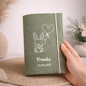 Könnte beinhalten: Ein hellgrüner Filz-Journal-Einband mit einem weißen Hasen, der einen herzförmigen Ballon hält. Der Name "Frauke" und das Datum "23.08.2025" sind weiß aufgedruckt. Ein weißes Gummiband sichert das Journal.