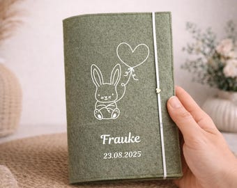 May include: A sage green felt journal cover with a white bunny holding a heart-shaped balloon. The name "Frauke" and the date "23.08.2025" are printed in white on the cover. A white elastic band secures the journal.