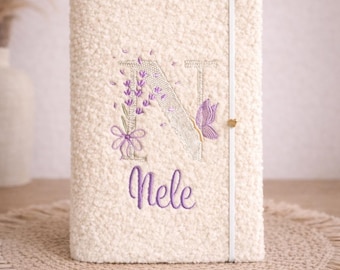 May include: Cream-colored journal with a textured cover. The letter "N" is embroidered in purple, adorned with lavender sprigs and a butterfly. The name "Nele" is embroidered below. A white elastic band secures the journal.