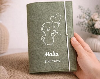 May include: A sage green felt journal with a white elastic closure. The cover features a white outline drawing of a penguin holding a heart-shaped balloon, with the name "Malia" and the date "31.01.2025" printed below.