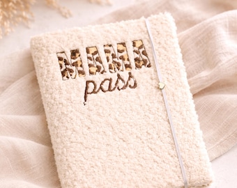 May include: Cream-colored, textured passport holder with the words "MAMA pass" embroidered on the front. The letters "MAMA" have a leopard print design. A thin, light-colored elastic band with a small heart charm secures the holder.