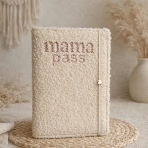 Könnte beinhalten: Ein cremefarbenes Notizbuch mit einer strukturierten Sherpa-Hülle, die mit dem gestickten Schriftzug "mama pass" in roségoldenem Garn versehen ist. Ein vertikales elastisches Verschlussband mit einem kleinen herzförmigen Metallanhänger.