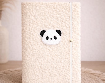 May include: Cream-colored, rectangular journal with a fuzzy, textured cover. A small panda face patch is centered on the front. A white elastic band with a gold-colored clasp secures the journal closed. The journal is sitting on a beige, round placemat.