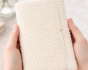 May include: Cream-colored notebook with a textured, plush cover. A white elastic band secures the notebook, with a small gold-colored accent. The notebook is held in two hands, suggesting a soft and tactile design.