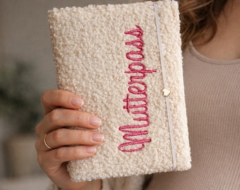 May include: Cream-colored, textured cover with the embroidered word "Mutterpass" in pink. A small gold heart charm is attached to the elastic closure. The cover is held in a hand.