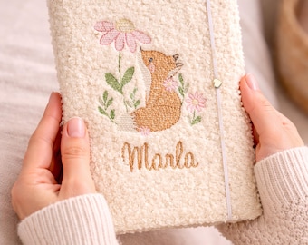 May include: Cream-colored journal with a textured cover, embroidered with a fox, pink flowers, and the name "Marla" in gold thread. A light-colored elastic band with a heart charm secures the journal.