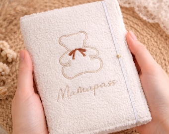 May include: Cream-colored, textured cover with an embroidered teddy bear and the word "Mamapass" in cursive. A white elastic band secures the cover. The cover is held in two hands.