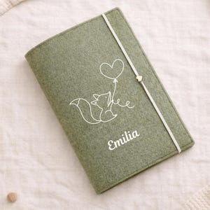 Könnte beinhalten: Salbeigrünes Filzjournal mit einer weißen Fuchsgrafik, die einen herzförmigen Ballon hält. Der Name "Emilia" ist in weißer Schrift aufgedruckt. Ein weißes Gummiband mit goldenem Herzverschluss sichert das Journal.