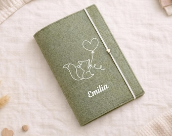 May include: Sage green felt journal with a white fox illustration holding a heart-shaped balloon. The name "Emilia" is printed in white script. A white elastic band with a gold heart closure secures the journal.