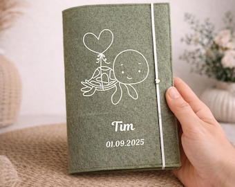 May include: Sage green felt journal with a white outline drawing of a turtle holding a heart-shaped balloon. The name "Tim" and the date "01.09.2025" are printed in white below the turtle. A white elastic band secures the journal.