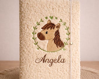 May include: Cream-colored, textured journal with an embroidered horse head in a green wreath. The name "Angela" is embroidered below the horse. A cream-colored elastic band secures the journal.