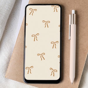 May include: A smartphone with a beige background and a pattern of brown bows. The phone is lying on a brown notebook with a pink pen next to it.