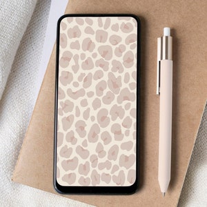 Pode incluir: Um smartphone com um papel de parede de leopardo bege e rosa na tela. O telefone está em cima de um caderno marrom com uma caneta rosa ao lado.