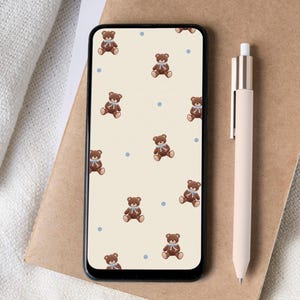 May include: A smartphone with a cream-colored background featuring a pattern of brown teddy bears with blue bows and dots. A light pink pen rests on a brown notebook next to the phone. A white cup is visible in the corner.
