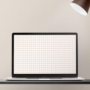 May include: A silver laptop with a black screen displaying a beige and white gingham pattern sits on a black surface. A brown desk lamp with a white interior is positioned above the laptop, casting light. The background is a neutral beige.