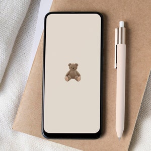 May include: A brown teddy bear sitting on a white background on a smartphone screen. The phone is on a brown notebook with a pen next to it.