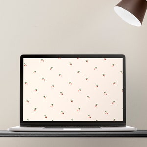 May include: A laptop computer with a white background and a repeating pattern of red cherries on the screen. The laptop is on a black desk with a brown lamp and a green plant in the background.