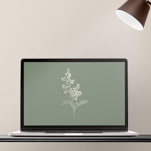 May include: A laptop displays a digital illustration of a white floral sprig on a sage green background. A brown desk lamp with a white interior is positioned above the laptop. The laptop sits on a black surface against a neutral wall.
