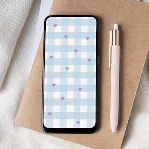 May include: A smartphone displaying a blue and white gingham pattern with purple heart motifs. A beige pen lies on a brown notebook, alongside a white cup on a coaster. The items are arranged on a white, textured surface.