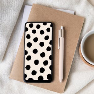 Könnte beinhalten: Ein Smartphone mit schwarzem Rahmen zeigt einen cremefarbenen Hintergrund mit schwarzen Punkten. Ein beiger Stift liegt neben dem Telefon auf einem braunen Notizbuch. Eine weiße Tasse und ein Untersetzer sind im Hintergrund.