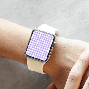 Puede incluir: Un reloj inteligente blanco con caja plateada y correa blanca se lleva en la muñeca. La esfera del reloj muestra un patrón de cuadros vichy morado y blanco. El fondo es gris neutro.