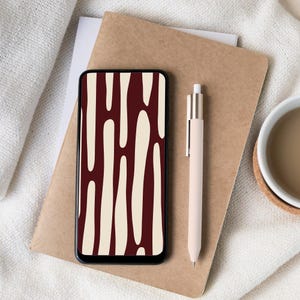 May include: A smartphone with a burgundy and cream abstract pattern is displayed on a brown notebook. A cream-coloured pen rests beside the phone. A white mug with a brown liquid sits on a coaster.