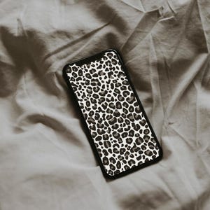 May include: A black smartphone case featuring a leopard print design in shades of gray, brown, and white. The case is rectangular and has rounded corners. The phone case is resting on a gray fabric surface.