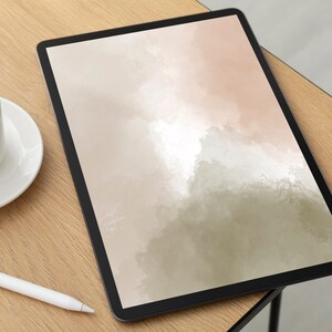 May include: A digital tablet with a black frame displays an abstract watercolor design in shades of beige, pink, and green. A white coffee cup and saucer sit to the left, with a white stylus pen nearby. The items are on a light wood surface.