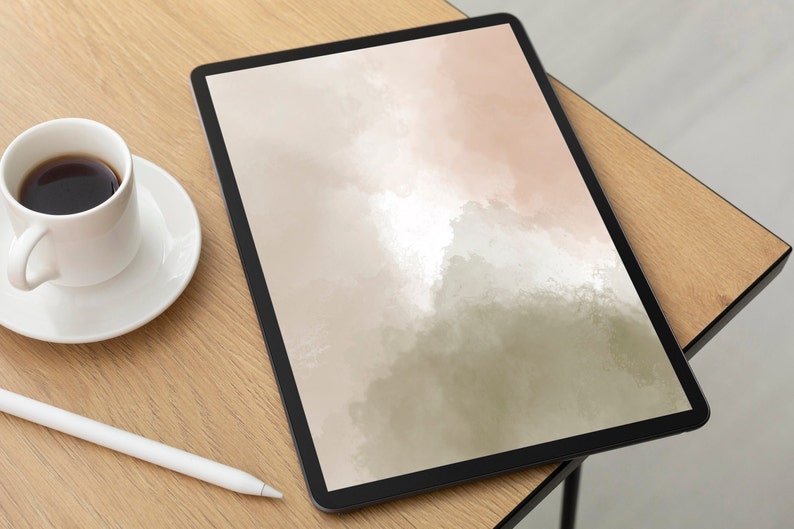 May include: A digital tablet with a black frame displays an abstract watercolor design in shades of beige, pink, and green. A white coffee cup and saucer sit to the left, with a white stylus pen nearby. The items are on a light wood surface.