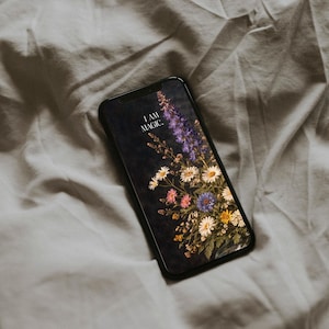 以下が含まれることがあります： 黒いスマートフォンには、「I AM MAGIC」というテキストが入った花の壁紙が表示されています。壁紙は、暗い背景に白い、ピンク、紫、黄色の花のブーケが特徴です。電話は灰色のしわのある布の上に置かれています。