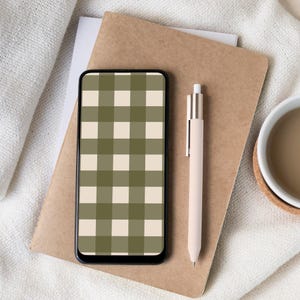Puede incluir: Un teléfono inteligente con un patrón de cuadros verdes y beige en la pantalla descansa sobre un cuaderno marrón. También hay un bolígrafo beige y una taza blanca con líquido marrón. Los objetos están dispuestos sobre una superficie blanca texturizada.