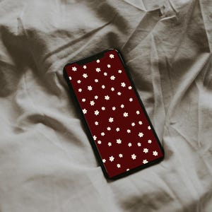 Könnte beinhalten: Ein Smartphone mit schwarzem Rahmen und einem weinroten Bildschirm, der ein Muster aus kleinen weißen Blumen zeigt. Das Telefon liegt auf einem zerknitterten, hellgrauen Stoff.