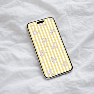 Puede incluir: Un smartphone con un patrón de rayas amarillas y blancas y pequeños acentos florales morados. El teléfono se muestra sobre una superficie blanca texturizada.