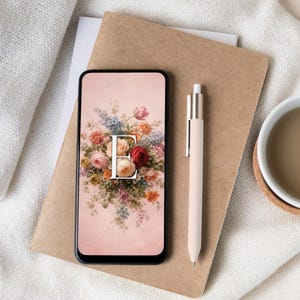 Op de afbeelding: Een smartphone toont een bloemenontwerp met de witte letter "E". Een lichtroze pen ligt naast de telefoon op een bruin notitieblok. Een witte mok met drank en een onderzetter staan op de achtergrond.