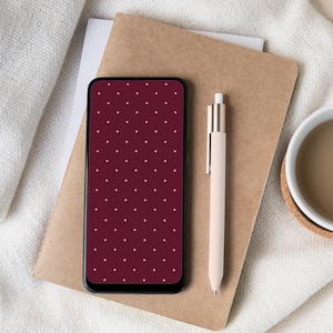 Könnte beinhalten: Ein Smartphone mit einem weinroten Bildschirm mit Polka-Dots liegt auf einem braunen Notizbuch. Ein beigefarbener Stift und eine weiße Tasse mit einem Getränk sind ebenfalls vorhanden. Die Artikel sind auf einer weißen Stoffoberfläche angeordnet.