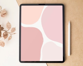 May include: A black tablet displays a digital art design with soft pink and peach-colored abstract shapes on a white background. A white stylus rests beside the tablet on a tan surface. Dried, gold-toned foliage is in the upper left corner.