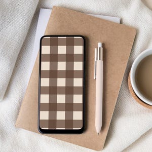 Puede incluir: Un teléfono inteligente con un patrón a cuadros marrón y beige en la pantalla descansa sobre un cuaderno marrón. También hay un bolígrafo beige claro y una taza blanca con líquido marrón. Los elementos están dispuestos sobre una tela blanca.
