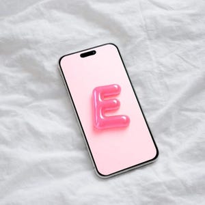May include: A smartphone with a pink screen featuring a glossy, bubblegum-pink letter "E". The phone has a silver frame and is set against a white, textured background. The letter "E" is the focal point.