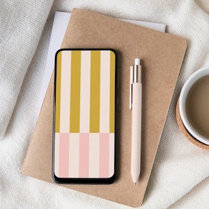 May include: A smartphone with a colourful striped pattern on the screen rests on a brown notebook. A light pink pen and a white mug with a brown liquid are also visible. The phone's screen features vertical stripes in mustard yellow, cream, and pink.