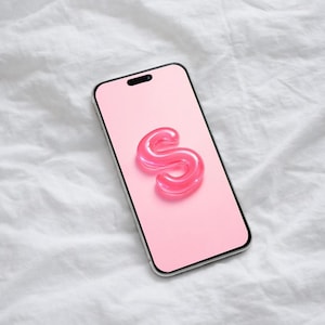 May include: A smartphone with a light pink screen displaying a glossy, three-dimensional letter S in a darker shade of pink. The device features a slim black frame and a centered camera cutout at the top of the display screen.