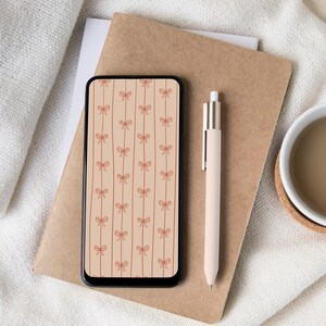 May include: A smartphone with a bow pattern on the screen rests on a brown notebook. A light pink pen and a cup of coffee are also visible. The bow pattern is in a repeating vertical design with a light brown background.