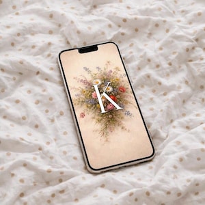 May include: A smartphone displays a floral wallpaper with the letter "K" in white. The wallpaper features a bouquet of daisies, red and pink flowers, and greenery. The phone is on a white fabric with tan polka dots.