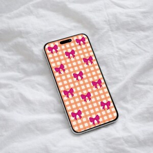 Puede incluir: Una funda de teléfono inteligente con un patrón repetido de lazos rosas sobre un fondo de cuadros de color melocotón y blanco. La funda del teléfono tiene un borde transparente y se muestra sobre una superficie blanca texturizada.