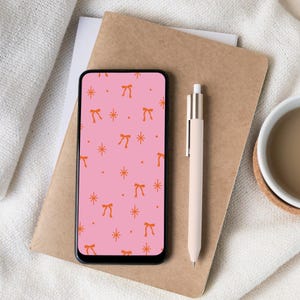 May include: A smartphone with a pink background featuring orange bows and stars, resting on a brown notebook. A beige pen and a cup of coffee are also present. The scene is set on a white, textured surface.