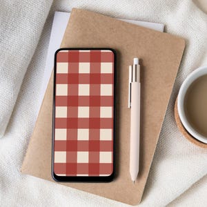 Puede incluir: Un teléfono inteligente con un patrón de cuadros rojos y crema en la pantalla descansa sobre un cuaderno marrón. También se ven un bolígrafo crema y una taza blanca con un líquido marrón. Los elementos están dispuestos sobre una superficie blanca texturizada.