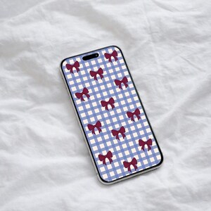 Puede incluir: Una funda de teléfono inteligente con un patrón de cuadros azul y blanco, adornada con ilustraciones repetidas de lazos burdeos. La funda tiene un borde transparente y se muestra sobre una superficie blanca texturizada.