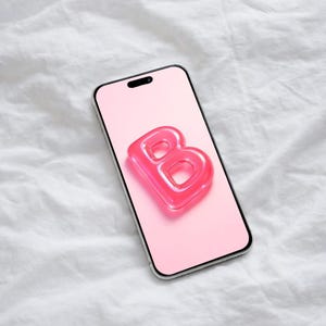 Könnte beinhalten: Ein Smartphone mit rosa Bildschirm zeigt den durchscheinenden, pinkfarbenen Buchstaben "B". Das Telefon hat einen silbernen Rahmen und liegt vor einem weißen, strukturierten Hintergrund. Der Buchstabe "B" scheint aus glänzendem Material zu bestehen.